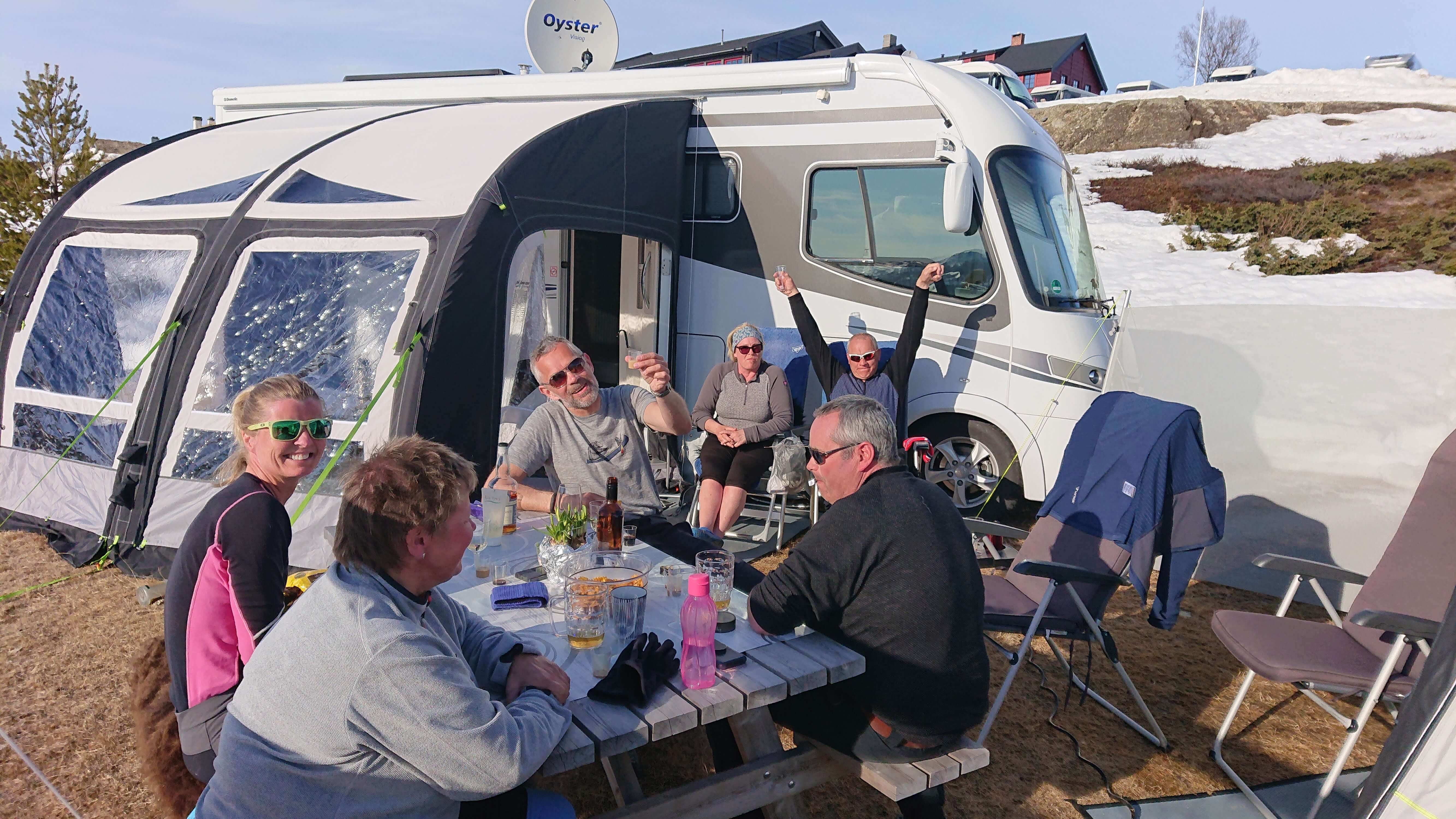 I bildet sitter en glad gjeng rundt et bord utendørs, med bobil, fortelt og snø i bakgrunnen.